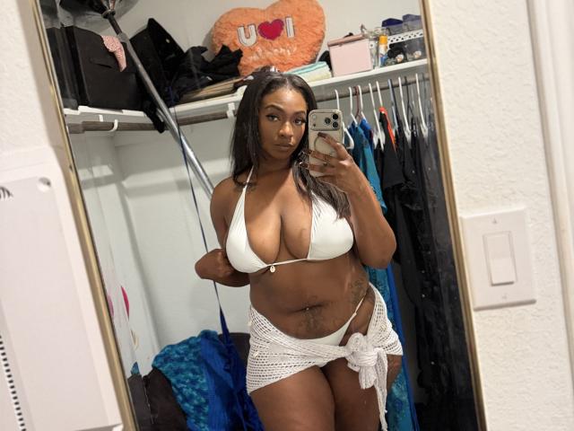 SunnyMochaQueen mit dunkler Haut, großen Brüsten in einem weißen Bikini-Oberteil, einem weißen Häkelrock, hält ein Telefon, Selfiemit Spiegel, langes schwarzes Haar, Tattoo am Bauch, Zimmer mit Kleidung..