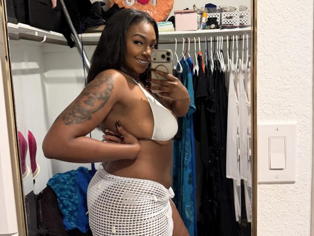 SunnyMochaQueen con la pelle scura, lunghi capelli neri, reggiseno bianco, che mostra il seno, gonna di rete bianca, tatuaggio sul braccio destro, sorridente, che si fa un selfie allo specchio nell'armadio..