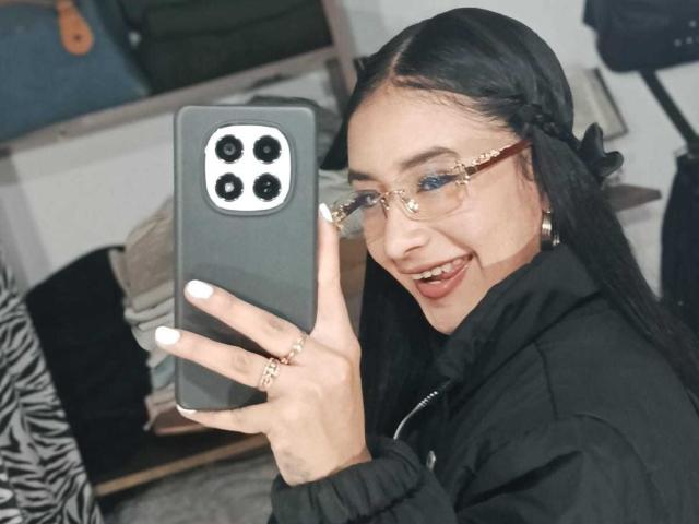 KatheLeclecr with long black hair, glasses, and a black headband, smiling while taking a selfie with a grey phone, wearing a black jacket and gold ring.
