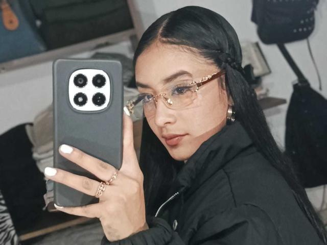 KatheLeclecr taking a selfie with a black iPhone, wearing glasses with gold frames, black jacket, long black hair, white nails, and gold rings, looking at the camera.