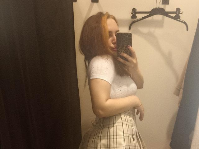 Maribelle Cherry mit roten Haaren, einem weißen Top mit kurzen Ärmeln und einem karierten Rock, macht ein Selfie vor einem Spiegel mit ihrem Handy und zeigt dabei ihre Brüste und ihr kurviges Hinterteil..