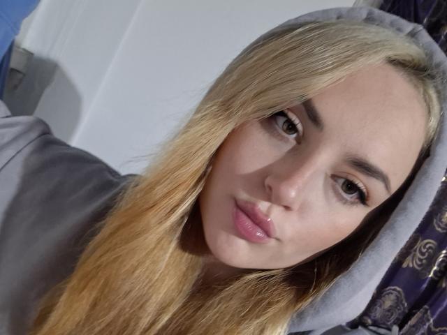 MaribelleCherry with blonde hair, brown eyes, pink glossy lips, wearing a grey hood, looking at the camera with a slight pout, soft light on her face.