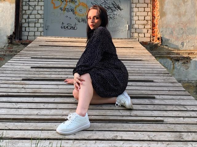 MaribelleCherry, hockend auf einer Holzplattform, trägt ein schwarzes Kleid mit weißen Punkten und weiße Turnschuhe, mit den Händen auf den Knien, und blickt über ihre Schulter..