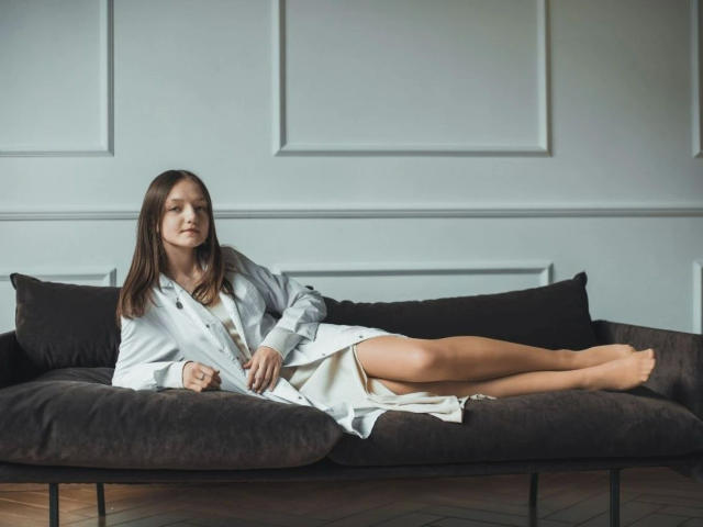 Keisyy-hot está deitada em um sofá marrom escuro, vestindo uma camisa e um vestido brancos, com o bumbum e os seios parcialmente visíveis, pernas cruzadas, e uma expressão relaxada e sonolenta..