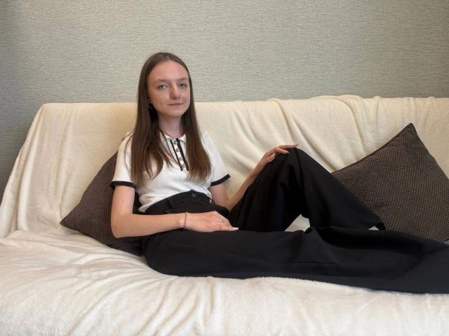 Keisyy, une jeune femme aux longs cheveux bruns, avec de petites seins, portant une chemise blanche et un pantalon noir, assise sur un canapé blanc avec des coussins marron, les jambes écartées, détendue, avec un léger sourire..