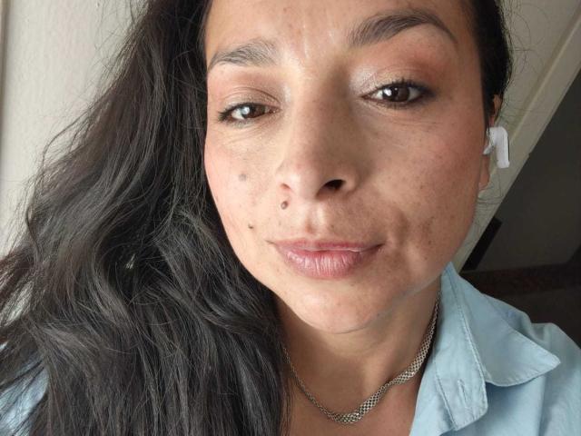 StangelarRivera has dark, wavy hair, brown eyes, light brown skin with freckles, small moles on her nose, and is wearing a light blue shirt with a silver chain.