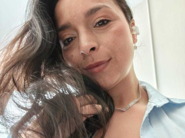 StangelarRivera with long brown hair, slight smile, wearing a light blue shirt and silver necklace, holding hair, brown eyes, small mole on left cheek.