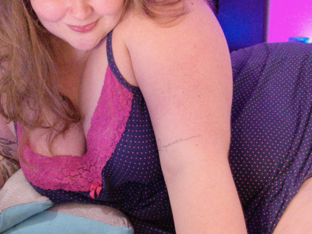 LilySweertx, com seus seios grandes, vestindo um vestido rosa com detalhes em renda e bolinhas, mostrando uma tatuagem no braço, sorrindo levemente, cabelo castanho, luz rosa ao fundo..