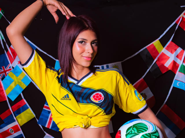 Danika Serov, con i capelli viola, indossa una maglia da calcio gialla, annodata davanti, tiene una palla da calcio, ci sono bandiere sullo sfondo, ha un braccio alzato, sta sorridendo e guarda la telecamera..