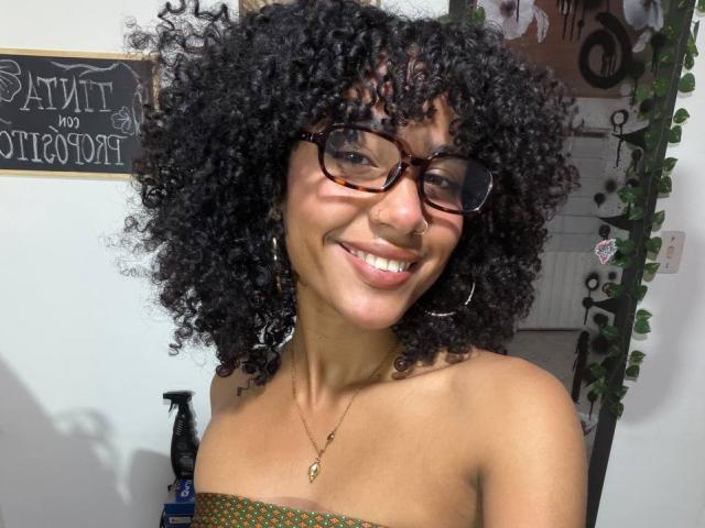 Cinthia Walker mit lockigem, schwarzem Haar, braunen, skorpionfarbenen Brillen, lächelnd, mit sichtbaren Zähnen, Reifenohrringen, einer goldenen Halskette, einem grünen, gemusterten Oberteil, gebräunter Haut und einem Nasenring..