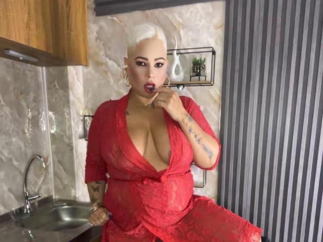 GianellaCox with platinum blonde hair, red lace dress, and large tits, sucking her finger in a kitchen. Her tattooed arm and gold earrings are visible.
