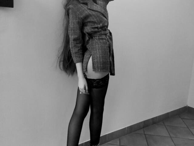 ElzaKomx indossa una giacca a quadri, ha i capelli lunghi, una maglia di pizzo nero e collant neri trasparenti, mostrando un piccolo sedere, ed è in piedi contro un muro..