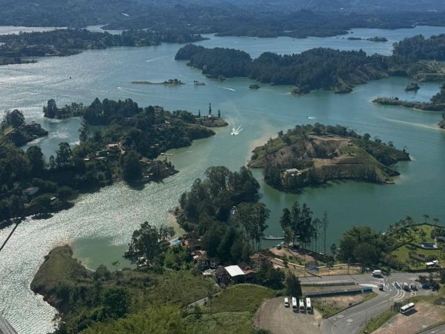 Vista aérea de un lago verde-azul con múltiples islas boscosas, un pequeño bote y una carretera con coches aparcados en la esquina inferior derecha. LilithDesire no es visible..