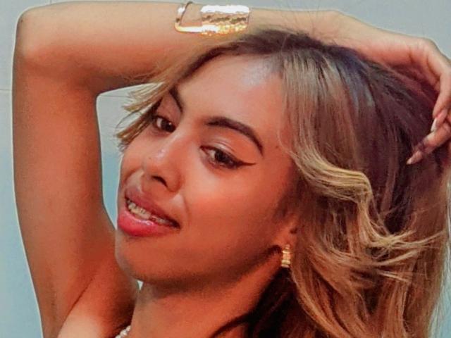 MissAniaSexy, com cabelo loiro ondulado, olhos castanhos e batom vermelho, usando uma pulseira dourada, olhando para o lado com a boca ligeiramente aberta, mostrando os dentes..