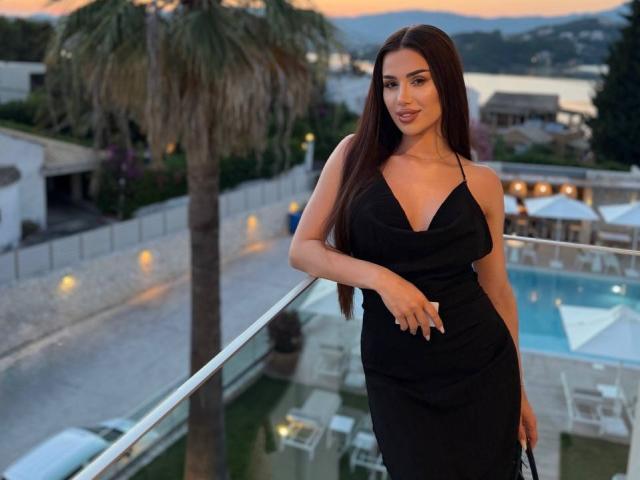 JennaDay está de pie en un balcón, vestida con un vestido negro, mostrando sus senos con una blusa ajustada, cabello castaño largo, sosteniendo un teléfono, fondo de atardecer, palmera, piscina, sillas blancas..