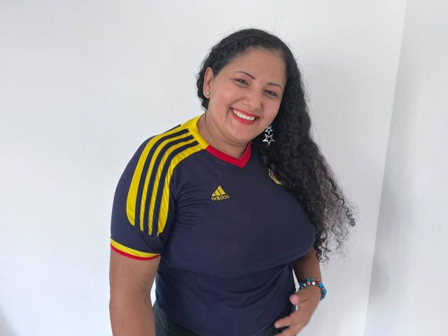 Genesis Montenegro, com cabelos pretos cacheados, sorrindo, vestindo uma camisa de futebol Adidas azul marinho com listras amarelas e detalhes vermelhos, exibindo seus seios grandes. Ela está usando brincos de estrela e um bracelete azul..
