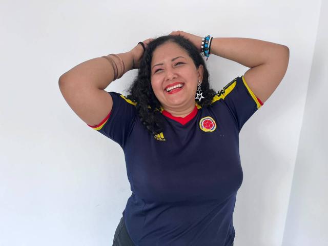 GenesisMontenegro mit lockigem, schwarzem Haar, lächelnd, mit den Händen im Haar, trägt ein dunkelblaues Fußballtrikot mit rotem Kragen, gelben Ärmeln und Armbändern, wobei ihre Brüste leicht durch das Trikot hindurch zu sehen sind..