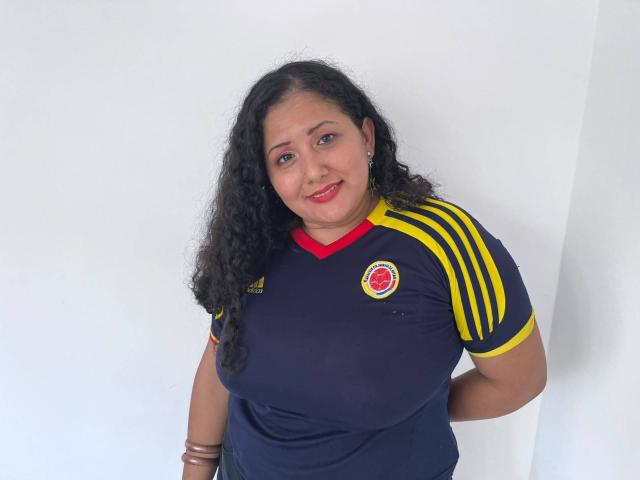 GenesisMontenegro mit lockigem, schwarzem Haar, rotem Lippenstift und dunkler Haut, trägt ein dunkelblaues Fußballtrikot mit gelben Streifen, lächelt in die Kamera und zeigt ihre kleinen Brüste und Arme..
