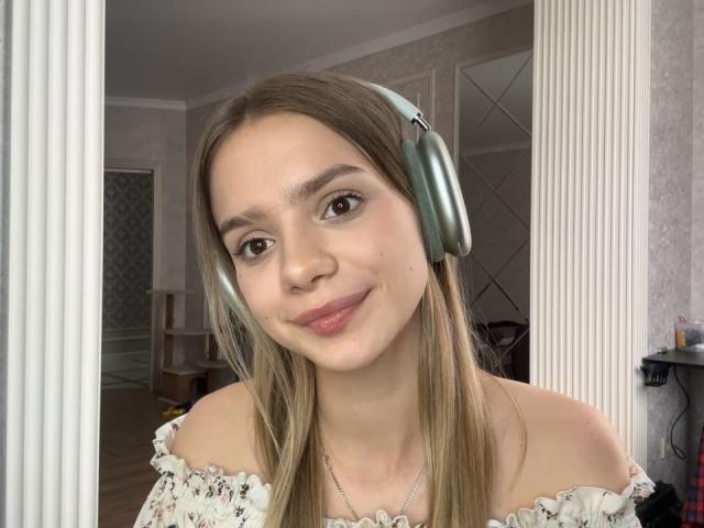 IrmaAncy with light brown hair, wearing metallic headphones, off-shoulder white top, smiling at camera, in a softly lit room. Boobs not visible. Ass not visible. Pussy not visible.