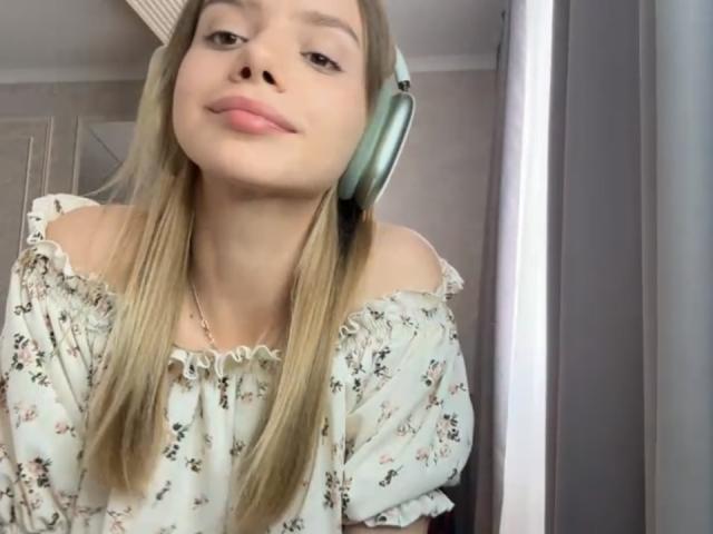 IrmaAncy with long blonde hair, off-shoulder floral top, and green headphones, smiling slightly, showing her cute nose and pink lips. Her tits are small, and she's cute.