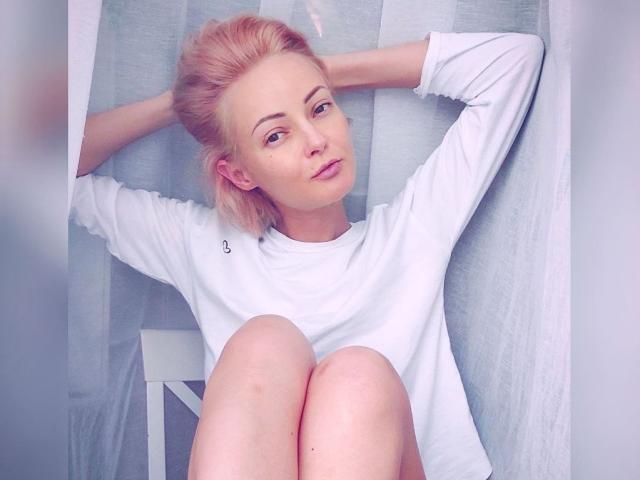 Una ragazza di nome Lollyypop, con i capelli rosa, indossa una camicia bianca a maniche lunghe, ha le braccia dietro la testa, le ginocchia sollevate e guarda la fotocamera con un leggero sorriso. Luce soffusa e naturale..