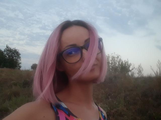 Lollypop, con cabello rosa, gafas y una blusa colorida, mirando hacia un lado con una ligera sonrisa. Sus glúteos no son visibles..