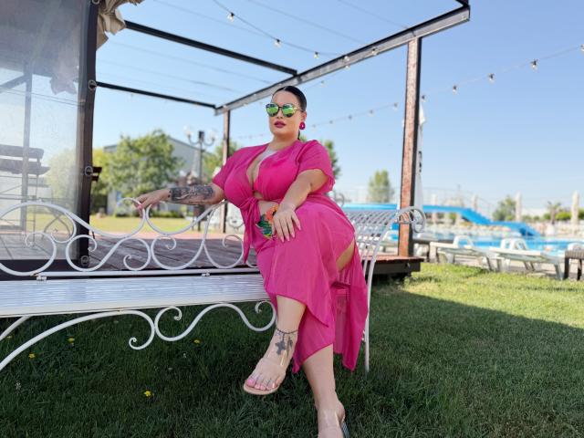 GeorgiaLerox, avec de grandes seins, une robe rose, des yeux verts, des boucles d'oreilles roses et des talons transparents, assise sur un banc blanc, avec un fond de prairie, un ciel clair, des guirlandes lumineuses et des tatouages sur son bras droit..