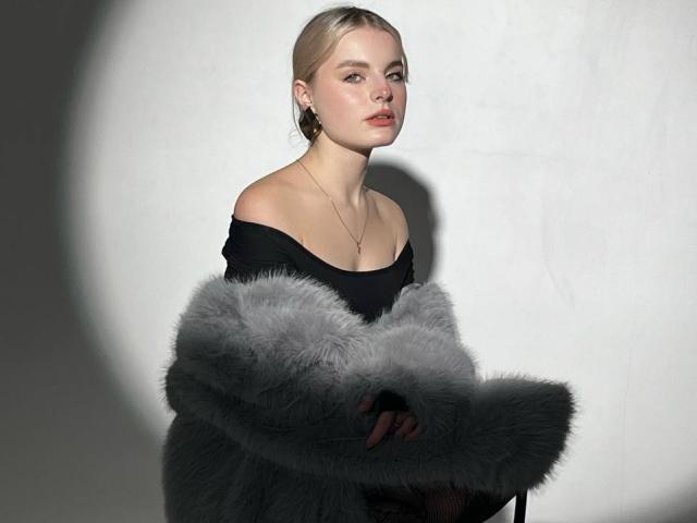AndreaFilmore, con capelli biondi, vestito nero con spalline scoperte, cappotto grigio in pelliccia, collana, viso in ombra, sguardo rivolto di lato, pelle chiara, trucco leggero, orecchini, luce soffusa, ambientazione in studio..