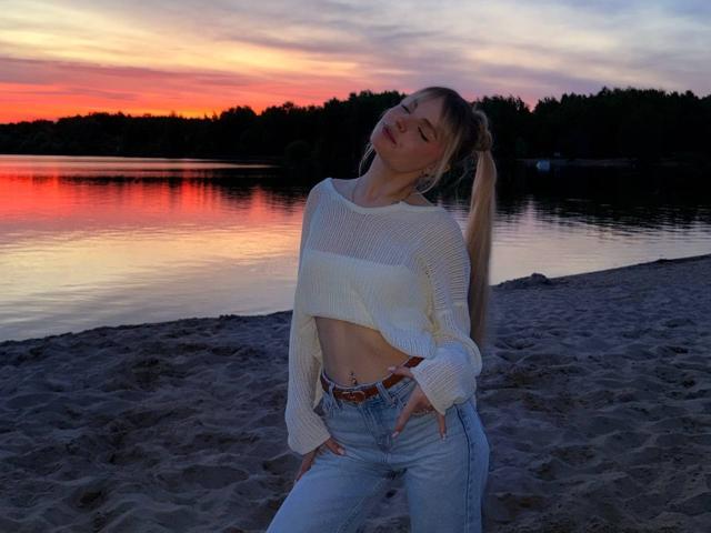 Andrea Filmore, con expresión de labios fruncidos, ojos cerrados, vistiendo una camiseta corta blanca, jeans azul claro, con las manos en los bolsillos, de pie en una playa arenosa, con un cielo al atardecer y agua reflectante en el fondo..