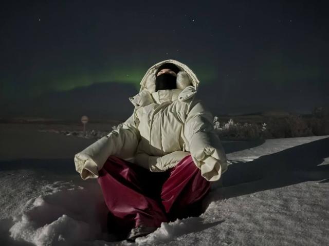 AndreaFilmore è seduta nella neve, indossa una giacca bianca imbottita e pantaloni rossi, con un cappuccio che le copre il viso, sotto un cielo verde illuminato dall'aurora boreale..