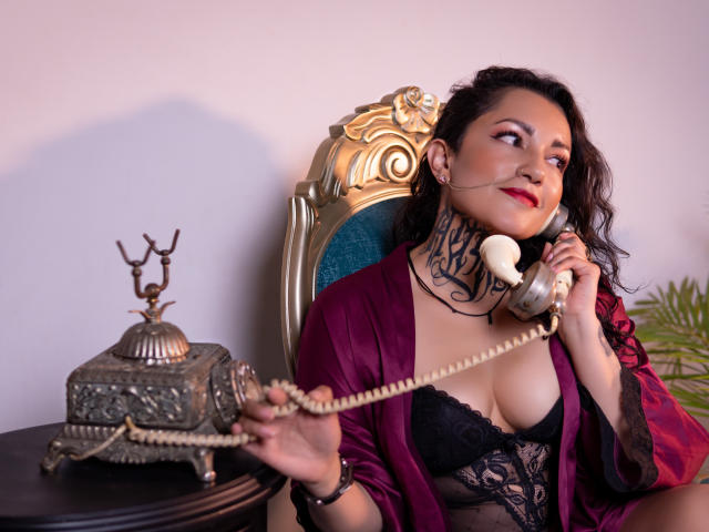 LorentGray, avec un soutien-gorge en dentelle noire, du rouge à lèvres rouge et une robe violette, tenant un téléphone vintage, montrant un tatouage sur son cou, assise sur une chaise dorée, à côté d'une boîte ornée..