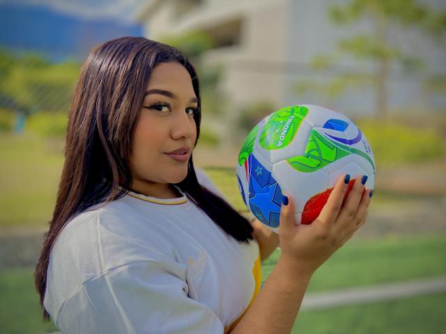 ValeryRiven holding a colorful soccer ball, wearing a white sports shirt, with long dark hair, and a slight smile, standing outdoors in bright sunlight.