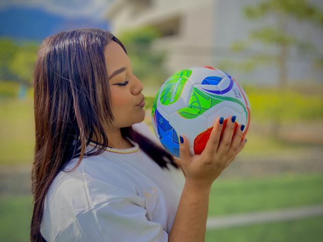 ValeryRiven mit glattem, braunem Haar, die einen bunten Fußball küsst, trägt ein weißes Hemd, hat blaue Nägel und hält den Ball vor einem sonnigen Hintergrund im Freien..