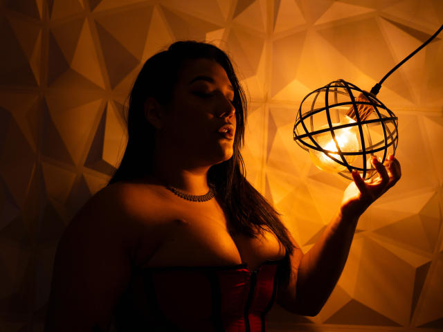 ValeryRiven with dark hair, wearing a red corset, holding a glowing orb lamp, casting warm light on her, showing her tits, in a dimly lit, geometric-patterned room.