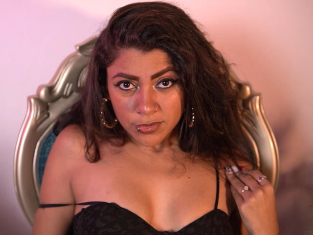 Natalia Cruz mit welligem, braunem Haar, die ein schwarzes Spitzen-BH trägt, sitzt in einem verzierten Stuhl, blickt direkt in die Kamera, hält an ihrem BH-Bügel und trägt einen Nasenring..