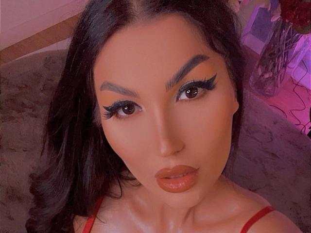 A ItsJessieBabe tem sobrancelhas grossas, cabelo preto longo e está usando batom vermelho com cílios pretos grossos. Ela tem uma expressão séria e está olhando para a câmera..