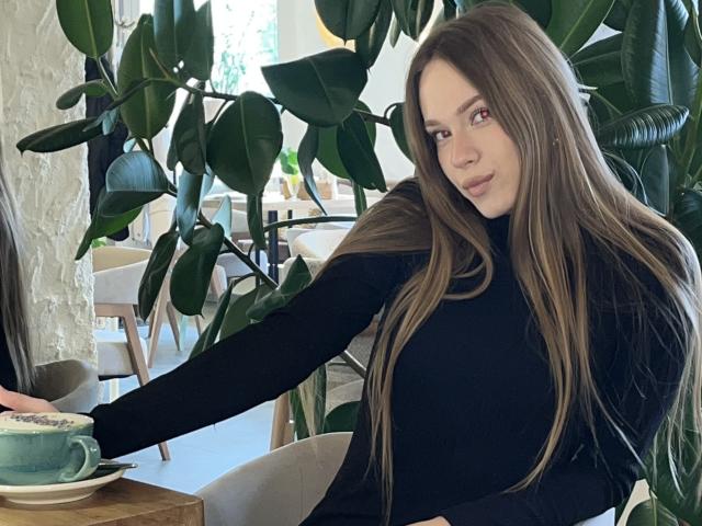 Kalyn Ivie, avec ses longs cheveux bruns, son pull noir à col roulé et son sourire charmant, tend la main vers une tasse verte posée sur une table en bois, entourée de plantes vertes à feuilles..