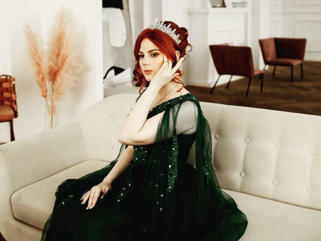 LilliamLechliter with red hair, wearing a green, sequined dress and silver tiara, sitting on a beige couch, hand on cheek, looking at camera, showing off her ass.
