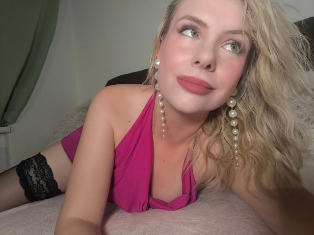 Étudiante - Jeune femme aux cheveux blonds, portant un haut rose à enfiler, des bas en dentelle noire montants jusqu'à la cuisse et des boucles d'oreilles de perles, allongée sur un lit rose, regardant sur le côté avec un sourire discret..