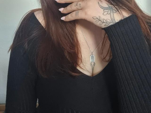 Valentina00-hot's hands with tattoos, black ribbed sweater, chest necklace, brown hair, showing cleavage. Her tits are slightly visible, nipples not shown. She's covering her mouth.