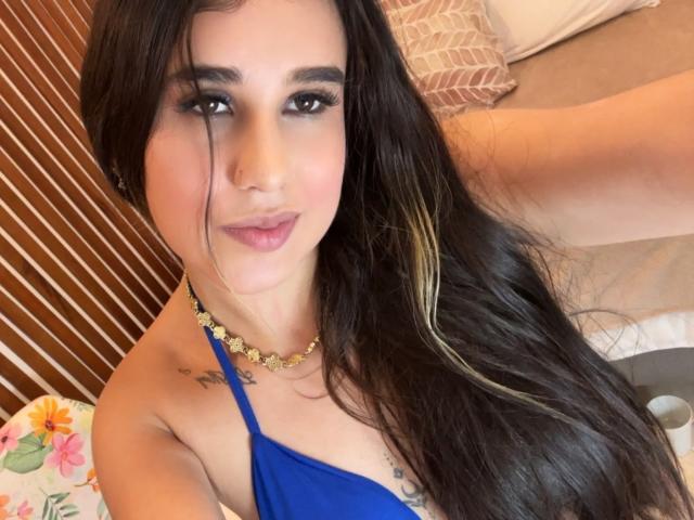 KatieDesirX, con lunghi capelli neri e riflessi biondi, indossa un top blu con spalline, una collana dorata e rossetto rosa, mostrando tatuaggi sul petto e un leggero sorriso..