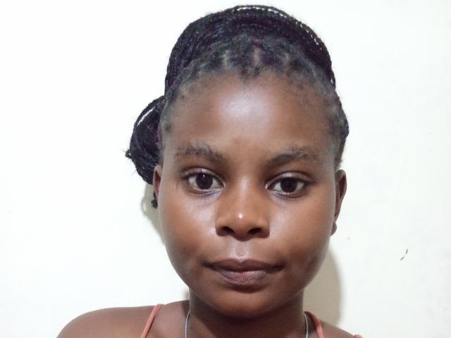 MinaAurora tem a pele marrom escura, olhos castanhos pequenos e cabelo preto trançado, preso para trás. Ela está vestindo uma blusa rosa, olhando diretamente para a câmera com uma expressão neutra..