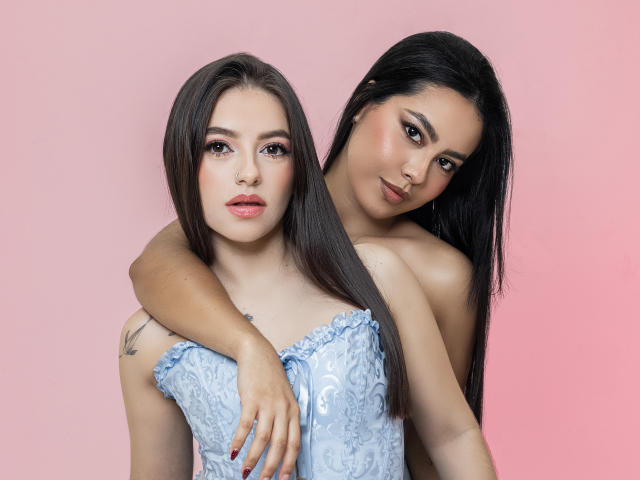 PaolaYKatty with long brown hair, blue lace-up corset, and tattoos, stands in front of another PaolaYKatty with black hair, arms around her, pink background.