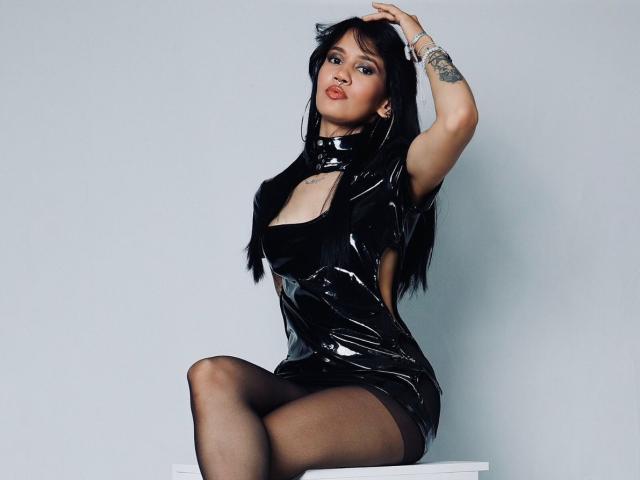 AryaAndSophie trägt ein glänzendes schwarzes Latexdress, sitzt mit verschränkten Beinen, trägt schwarze Strumpfhosen, hat einen tätowierten Arm, langes schwarzes Haar, voller Lippen, eine Hand im Haar, eine sexy Pose..