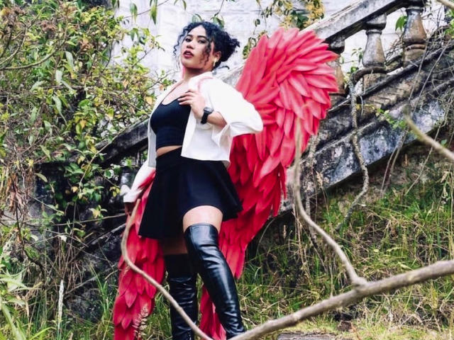 SophieSonyas com asas vermelhas, vestido preto, jaqueta branca e botas pretas que vão até a coxa, em pé na grama, tem cabelo preto encaracolado, maquiagem escura e um relógio. Seios visíveis, aparência sexy..