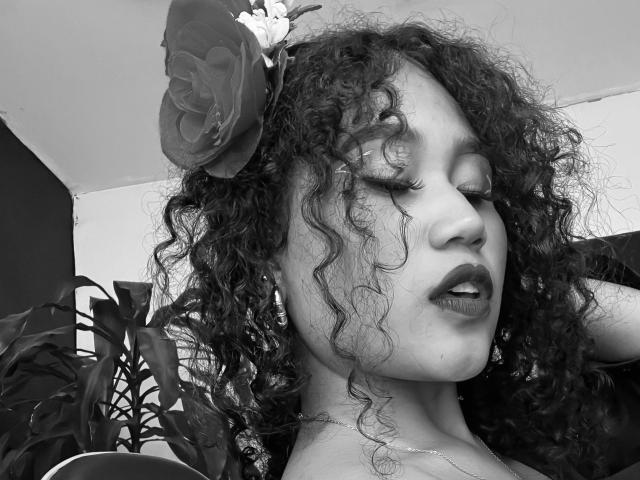 SophieSonyas tiene el pelo oscuro y rizado, un sombrero con flores y los ojos cerrados, con lápiz labial oscuro. Su nariz está ligeramente levantada y lleva un collar. Fotografía en blanco y negro..