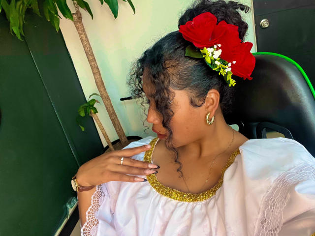 SophieSonyas com cabelo preto encaracolado, adorno de flor vermelha no cabelo, brincos de ouro, vestido branco com detalhes em renda dourada, anel no dedo, olhando para o lado, sentada em uma cadeira preta..