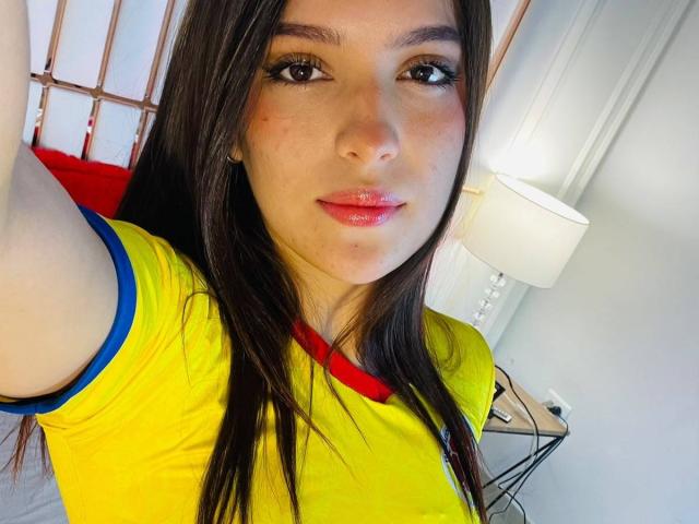 Katarina Novak, met lang bruin haar, draagt een geel shirt met rode en blauwe accenten, heeft roze lippen en maakt een selfie, waarbij haar gezicht en arm te zien zijn..