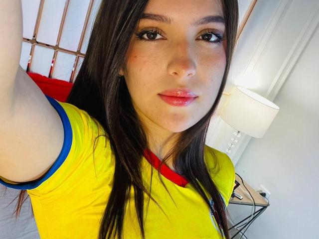 KatarinaNovak with long brown hair, wearing a yellow shirt with blue and red trim, has glossy pink lips and a slightly flushed face, taking a selfie in a bright room.