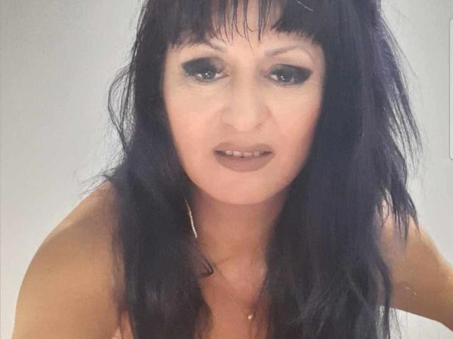 YummyTitss has long, dark, messy hair, black eyeliner, and brown lipstick. She's looking at the camera with a slight, open-mouthed smile, showing her teeth. Her tits are visible.